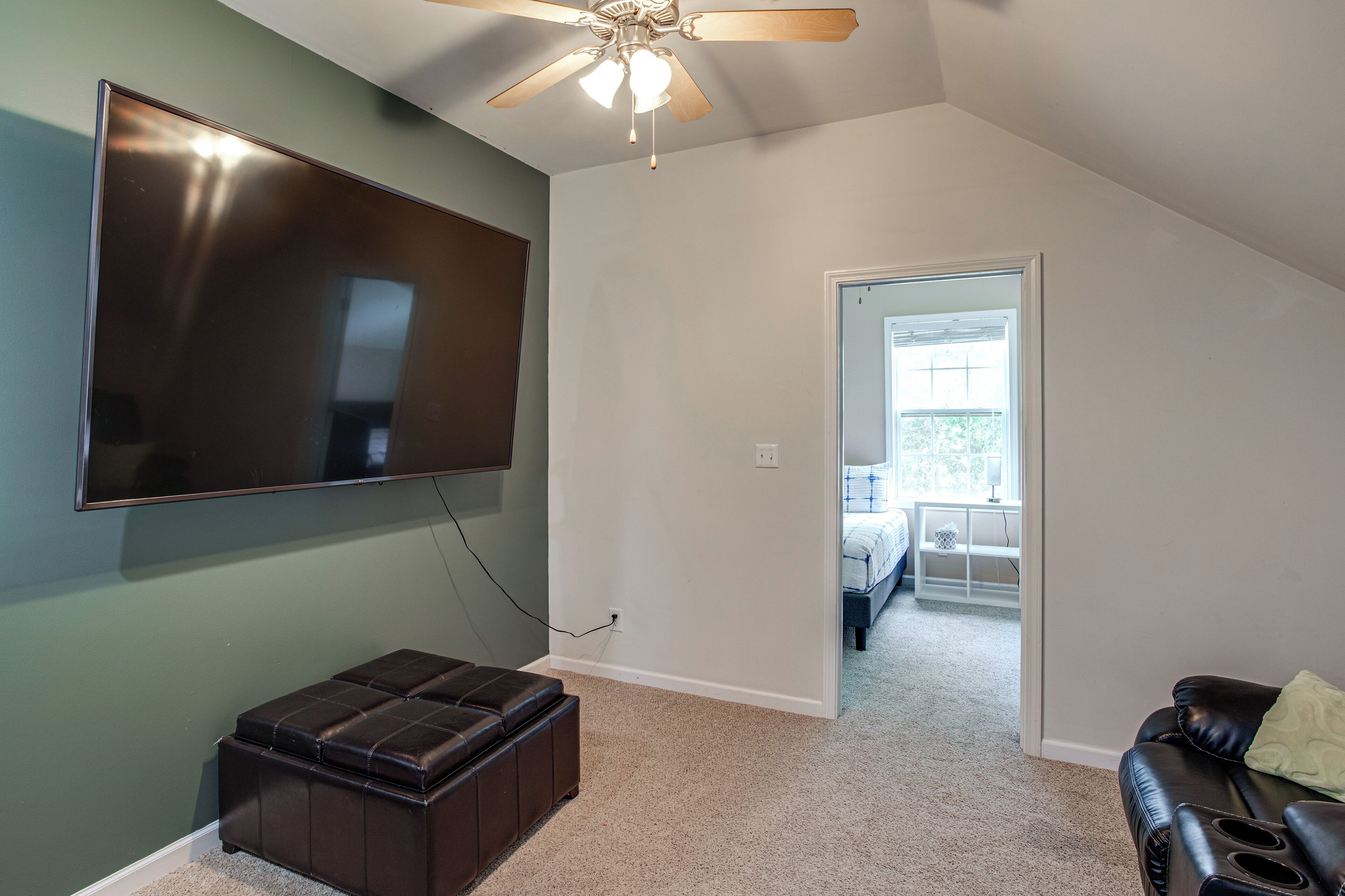 A room with a flat-screen TV mounted on the wall, a ceiling fan, a dark brown ottoman, and a doorway leading to another room.