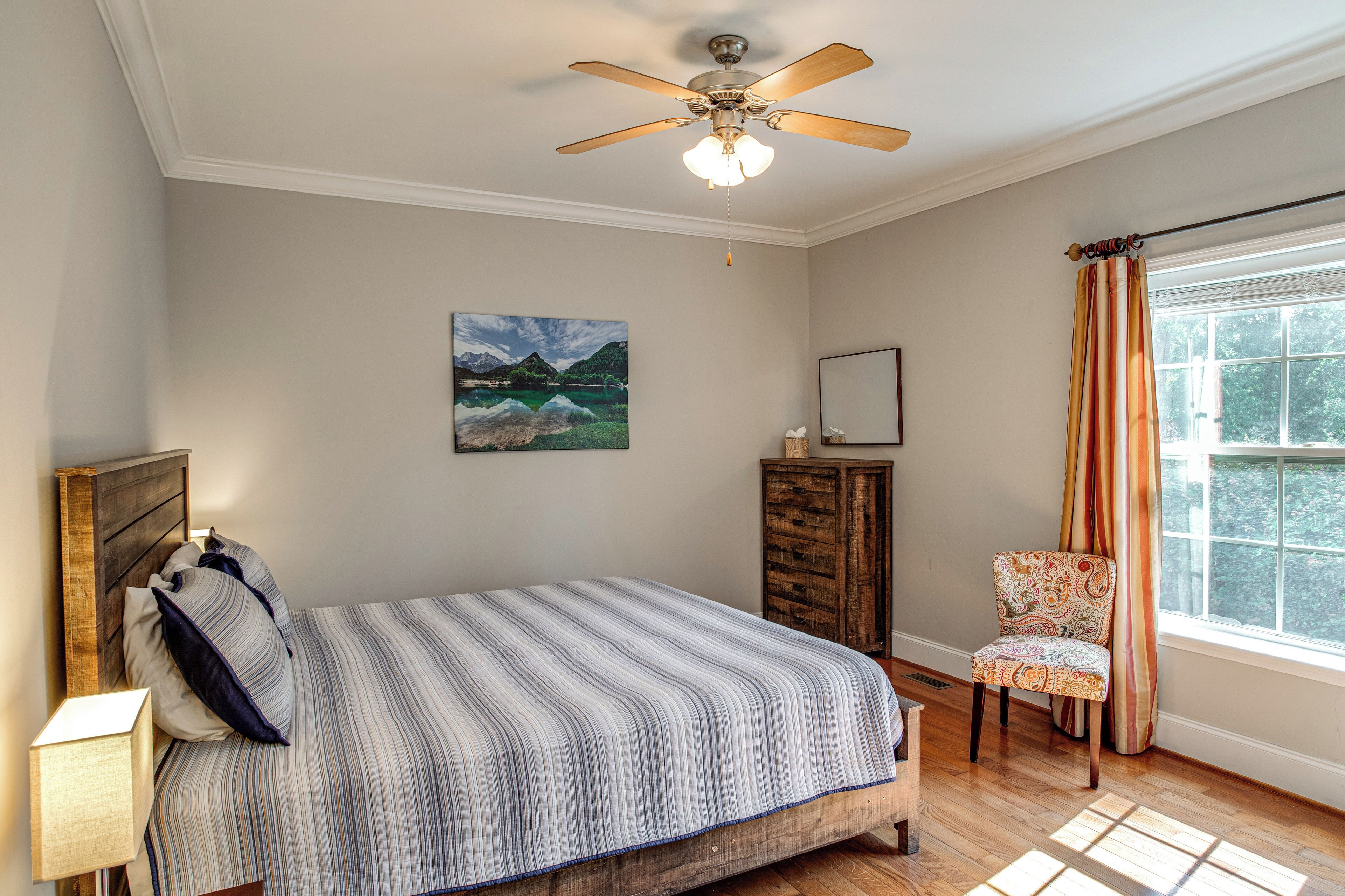 A bedroom with a bed, a ceiling fan, a painting, a dresser, a chair, and a window with curtains.