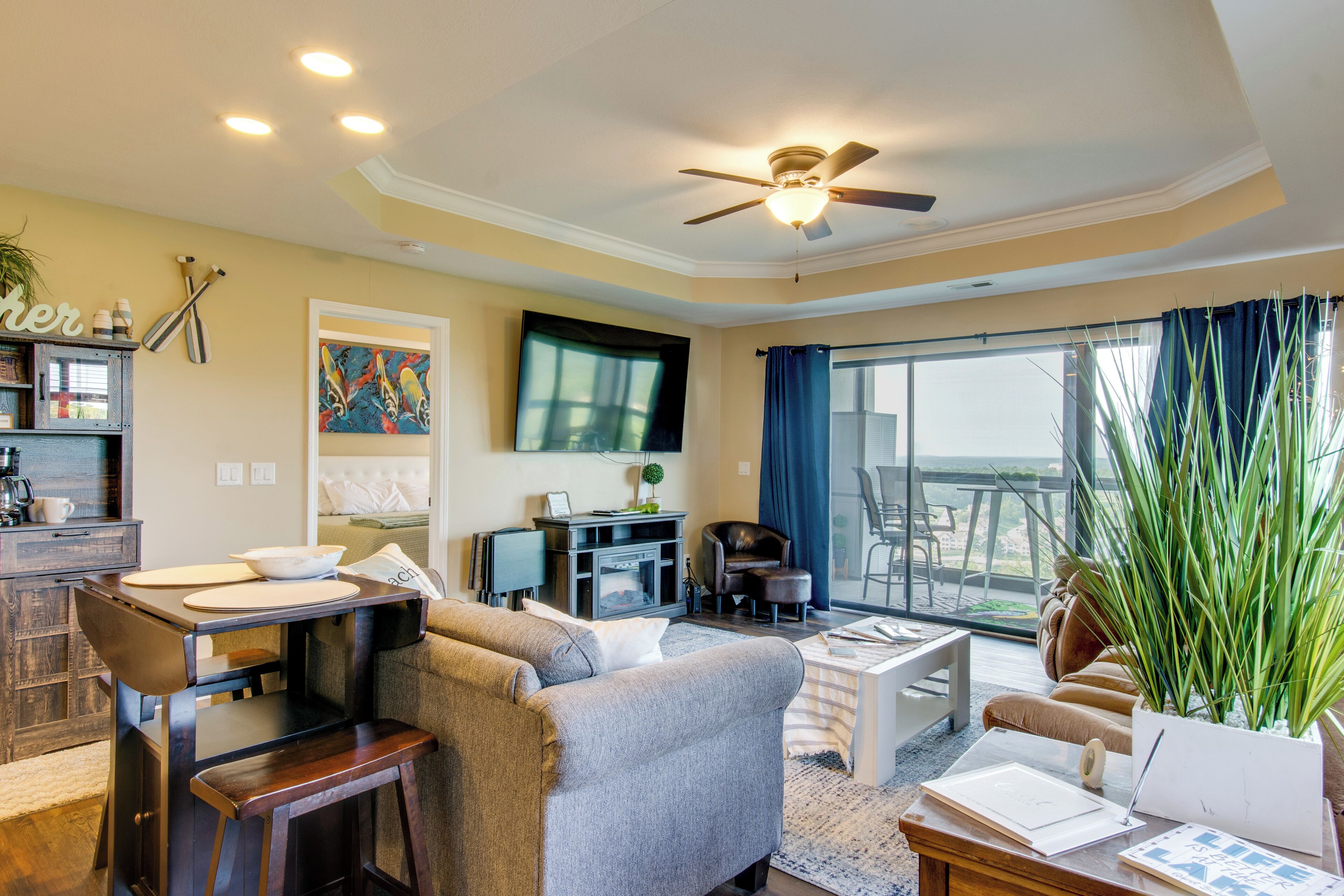 A living room with a sofa, coffee table, and a dining area with a table and chairs. There is a television mounted on the wall and a ceiling fan.