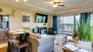 A living room with a sofa, coffee table, and a dining area with a table and chairs. There is a television mounted on the wall and a ceiling fan. - 'the Wake Zone' Condo Near Lake of the Ozarks (Lake Ozark)