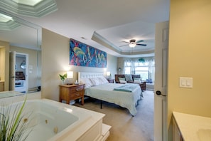 A bedroom with a large bed, a nightstand, a dresser, a ceiling fan, a window with curtains, and a bathroom with a bathtub. - 'the Wake Zone' Condo Near Lake of the Ozarks (Lake Ozark)
