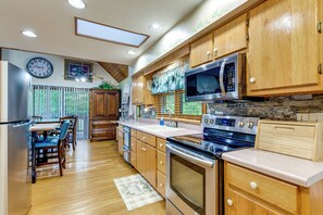 A kitchen with wooden cabinets, stainless steel appliances, a dining area with a table and chairs, and a clock on the wall. - Creekside Maggie Valley Home w/ Mtn-view Creek-vi (Waynesville)