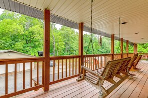 A covered porch with wooden railings, a swing, and a view of a green forest. - Creekside Maggie Valley Home w/ Mtn-view Creek-vi (Waynesville)