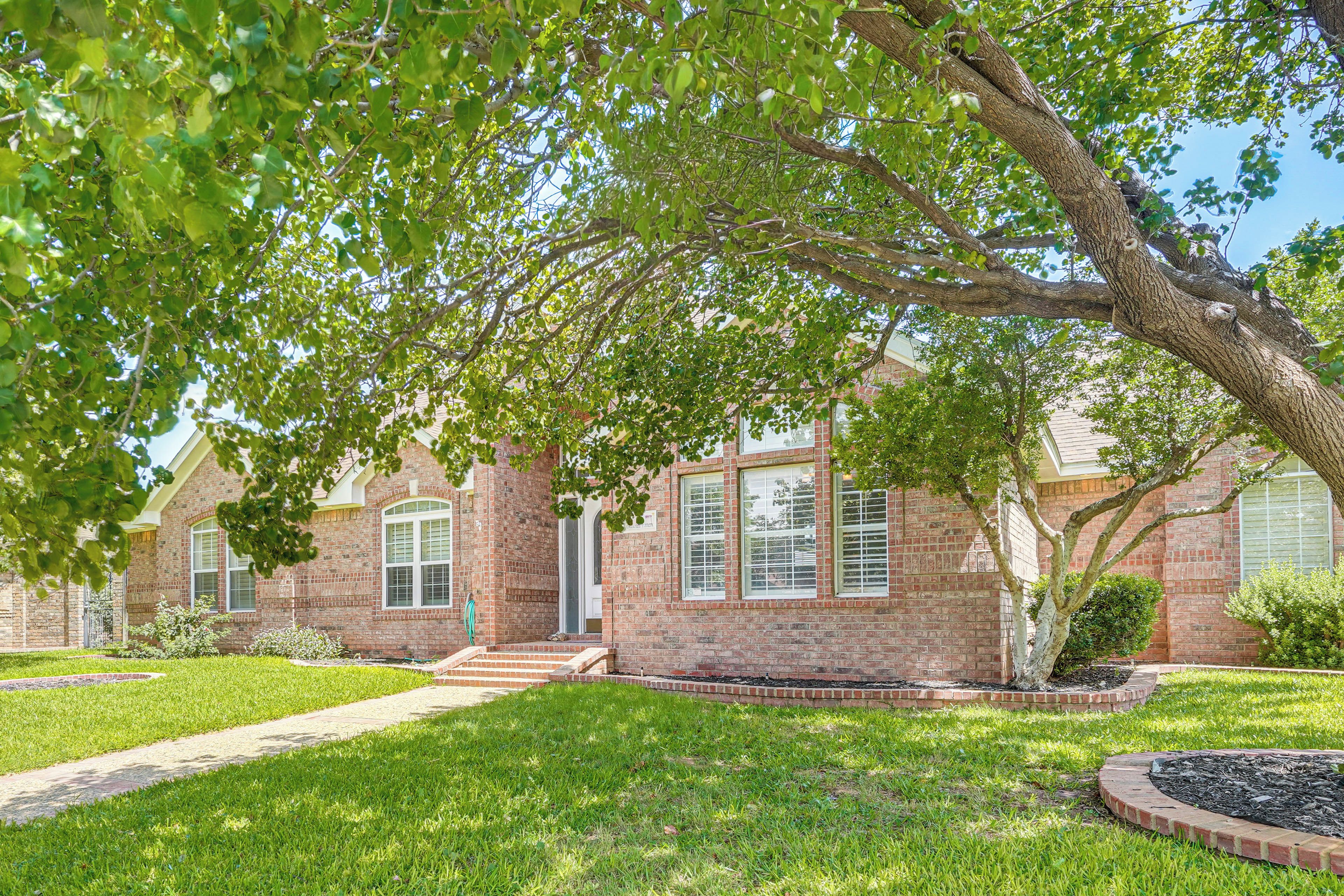 A brick house with a front yard, a pathway leading to the entrance, and a large tree providing shade.