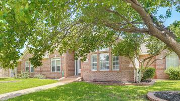 A brick house with a front yard, a pathway leading to the entrance, and a large tree providing shade.