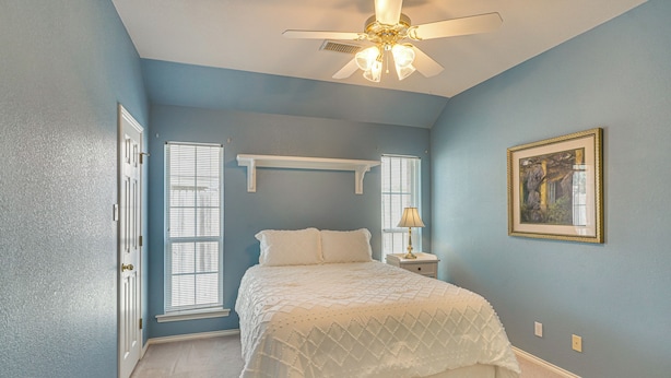 A bedroom with a bed, a ceiling fan, a nightstand, a lamp, a window with blinds, and a framed picture on the wall.