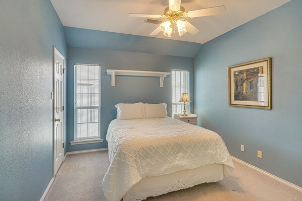 A bedroom with a bed, a ceiling fan, a nightstand, a lamp, a window with blinds, and a framed picture on the wall.