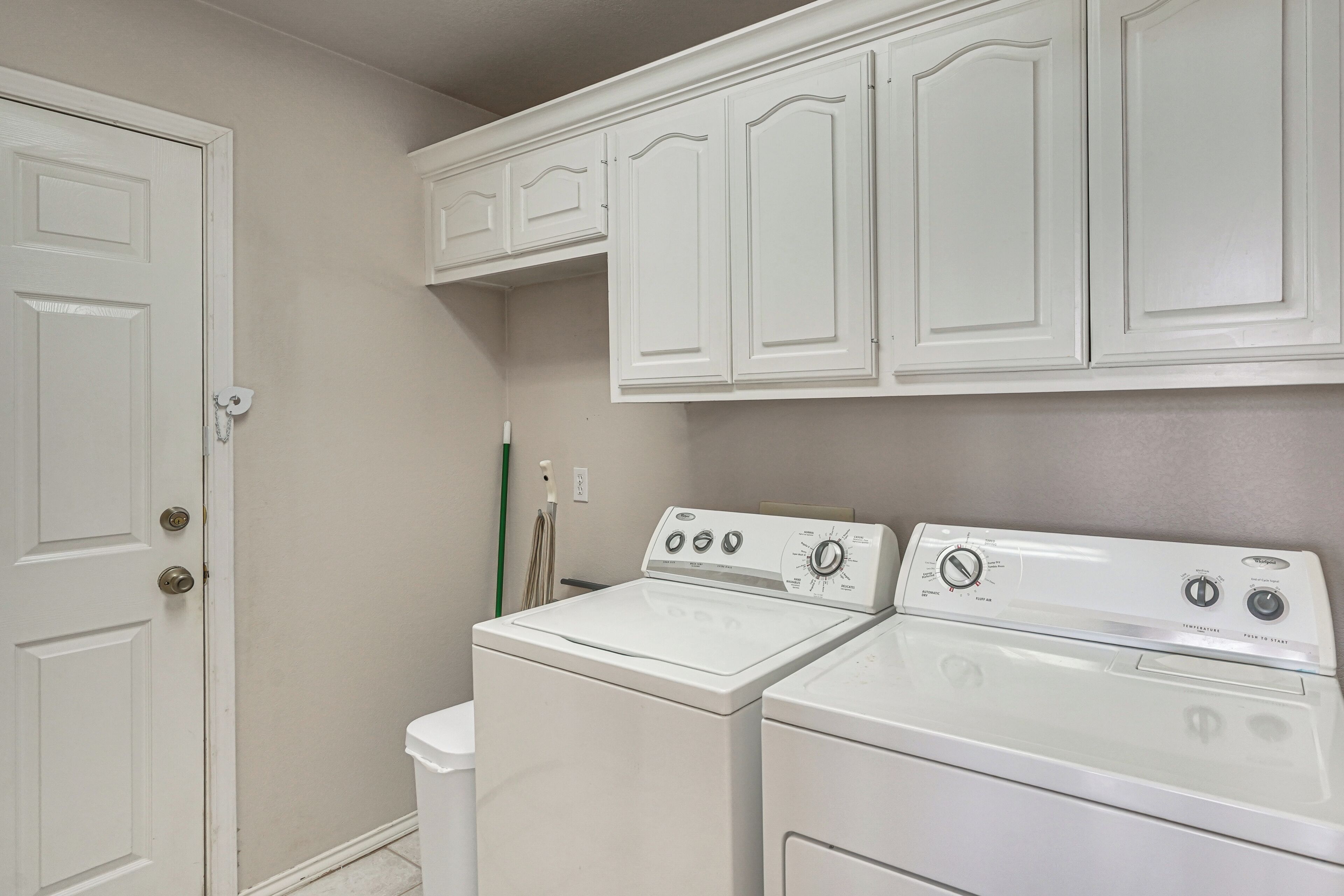 A laundry room with a front-loading washer and dryer, a door with a key lock, and a mop leaning against the wall.