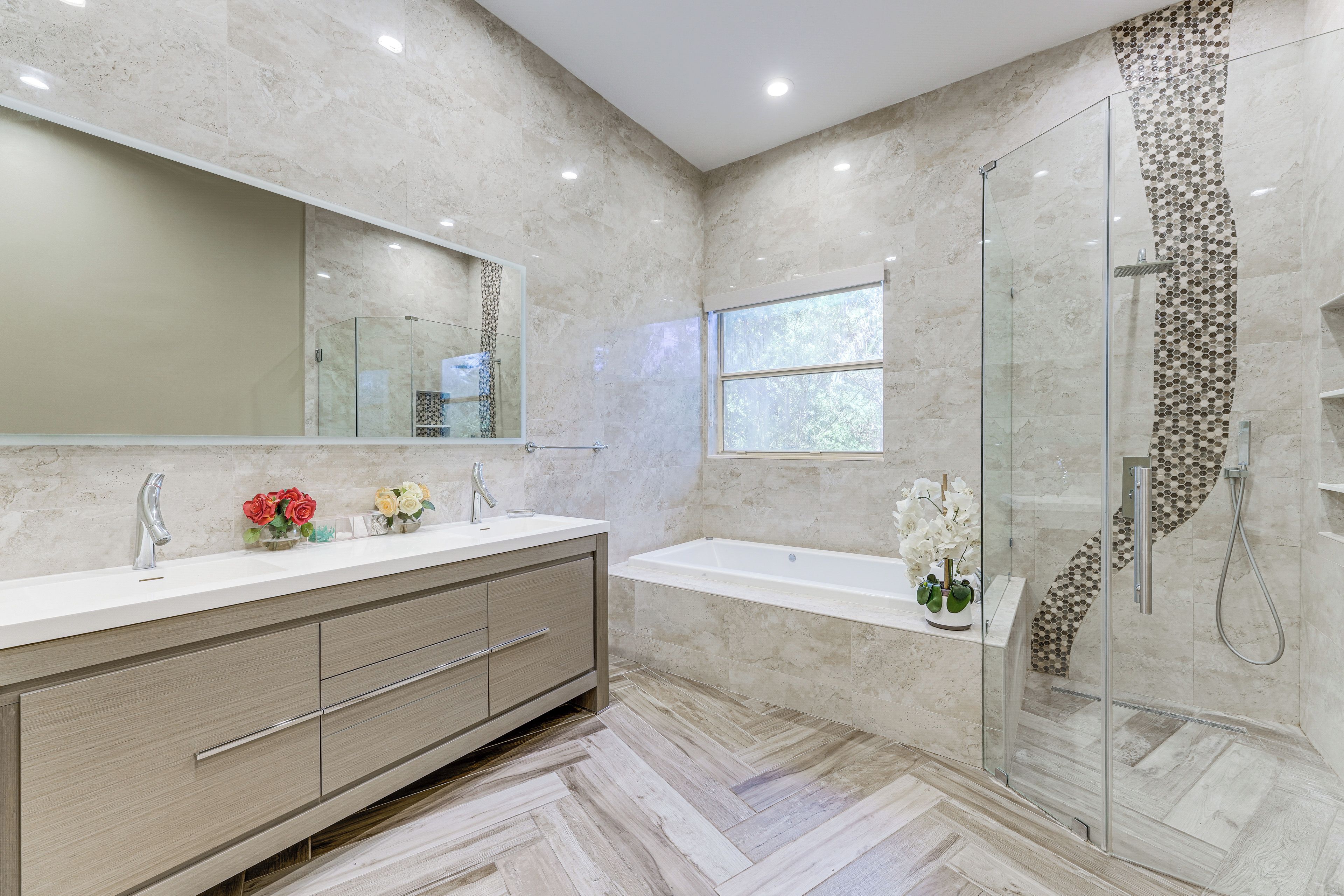 A modern bathroom with a glass shower enclosure, a large mirror, and a vanity with a sink and multiple flower arrangements.
