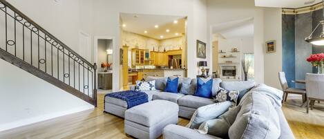 A spacious living room with a grey sectional sofa, a wooden staircase, and a kitchen in the background.