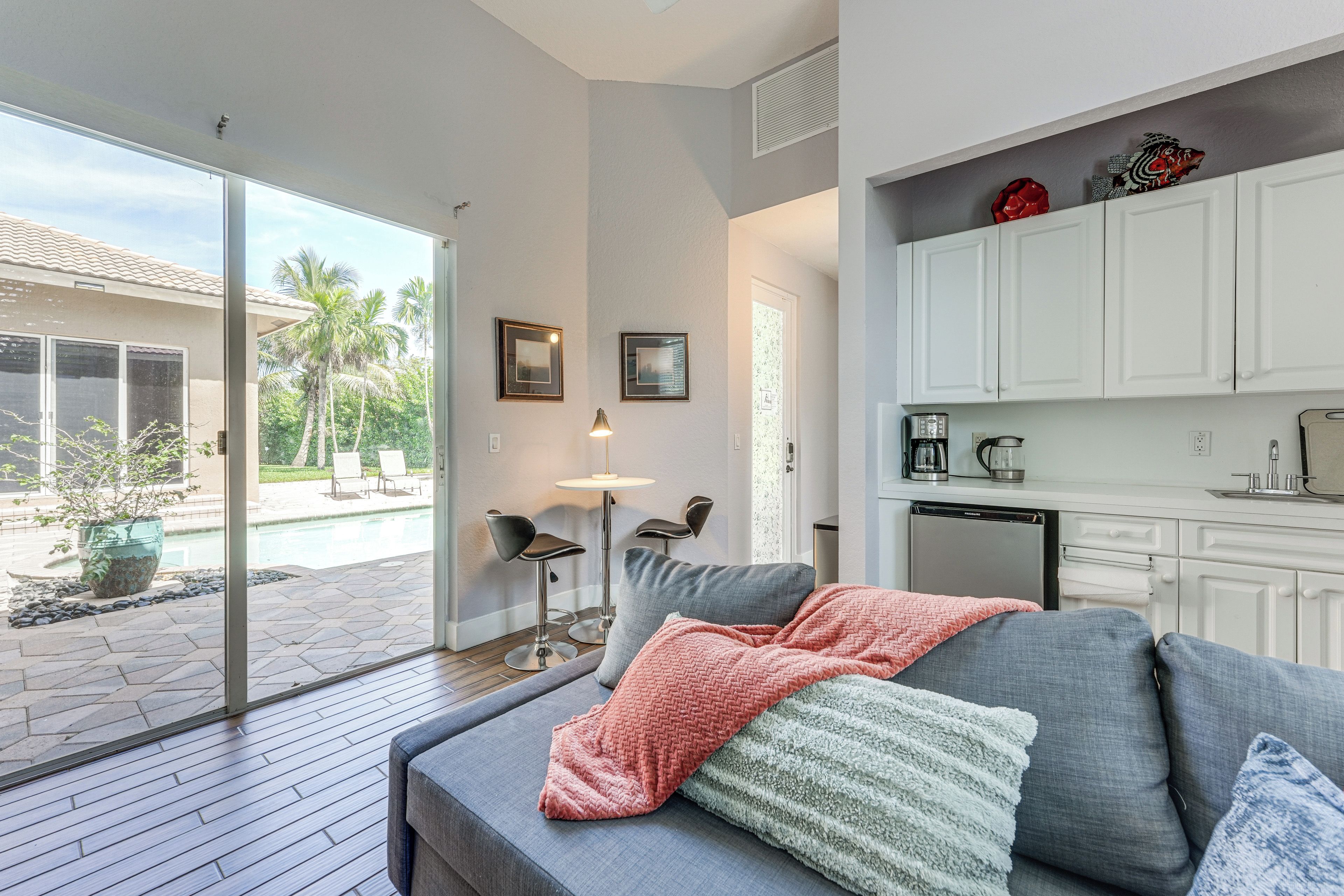 A modern living room with a grey sofa, a coffee table, and a view of a pool and palm trees outside.