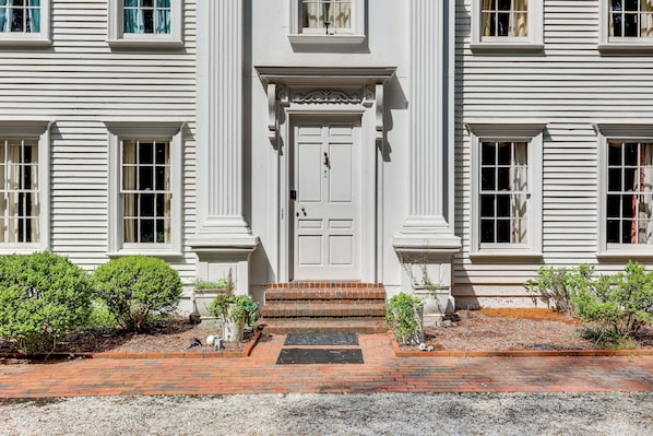 A two-story building with a symmetrical facade, a central entrance with a double door, and a brick pathway leading to the entrance.