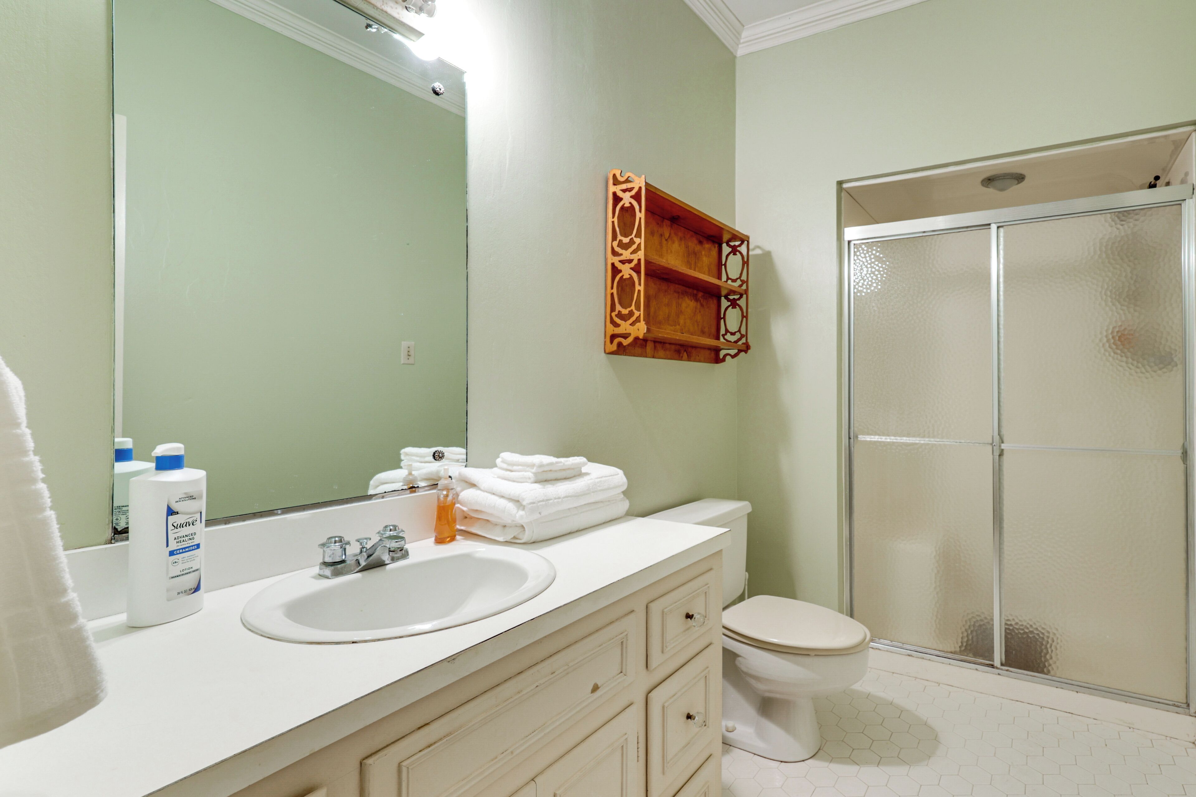 A bathroom with a white sink, mirror, and toilet. There is a shower with a glass door and a wooden shelf above the sink.