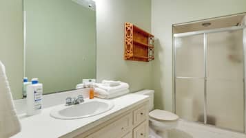 A bathroom with a white sink, mirror, and toilet. There is a shower with a glass door and a wooden shelf above the sink.