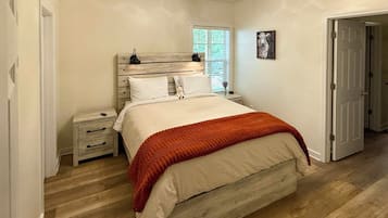 A bedroom with a wooden headboard, a bed with white bedding and an orange throw, a nightstand with a lamp, and a window with blinds.