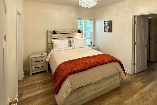 A bedroom with a wooden headboard, a bed with white bedding and an orange throw, a nightstand with a lamp, and a window with blinds.