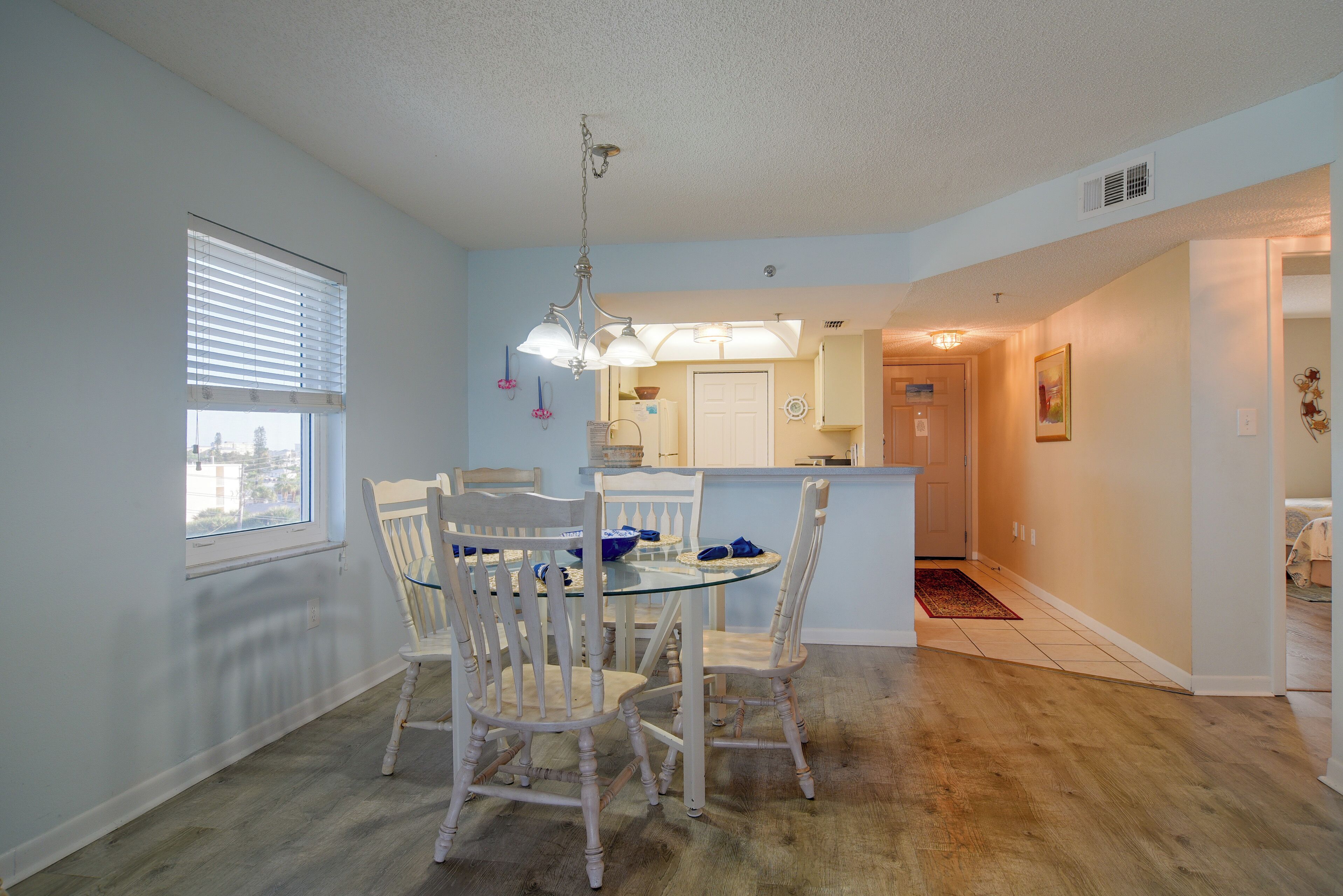 A dining area with a glass table and white chairs, a kitchen in the background, and a hallway leading to another room.