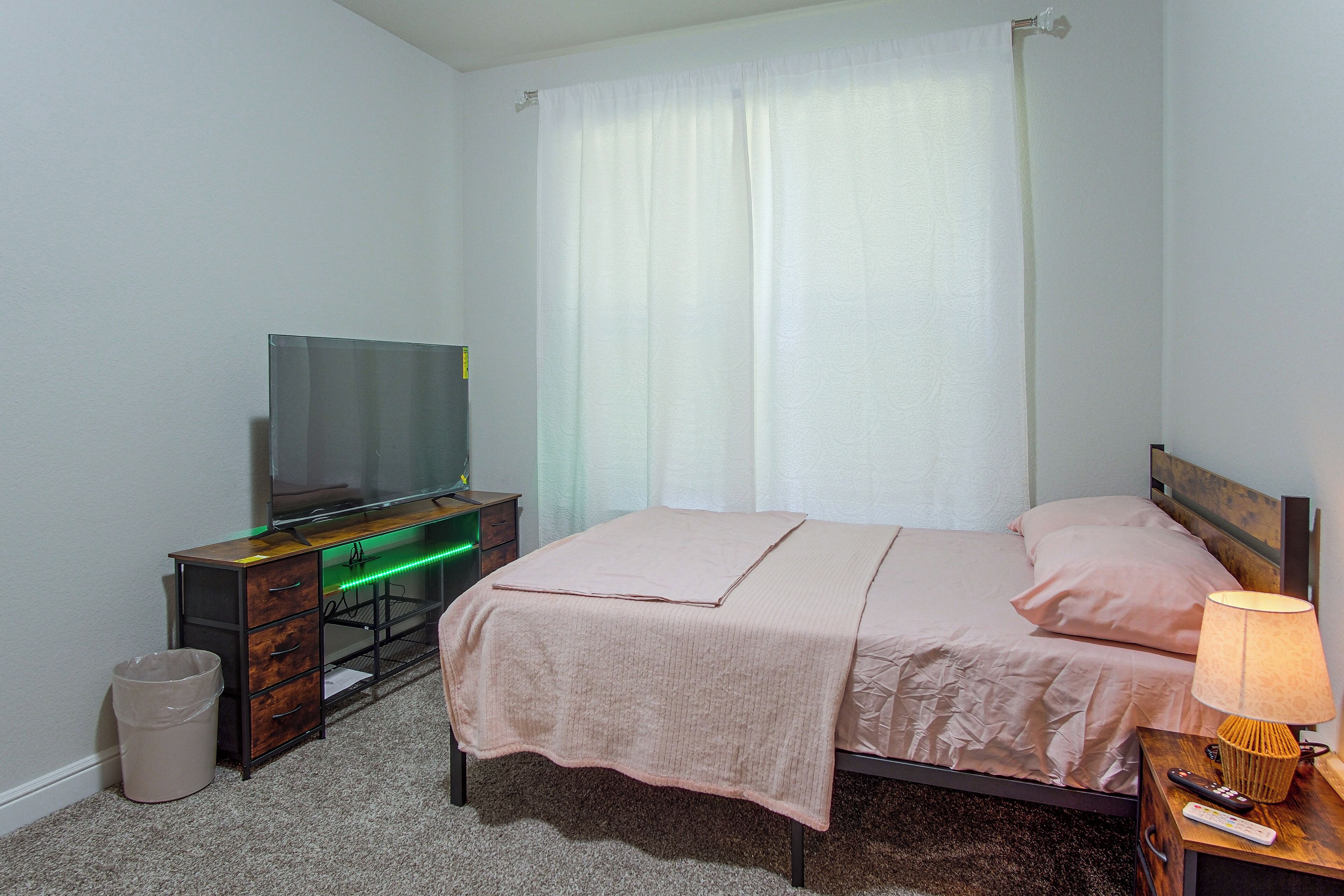 A hotel room with a bed, a television on a wooden dresser, a lamp, and a trash bin.