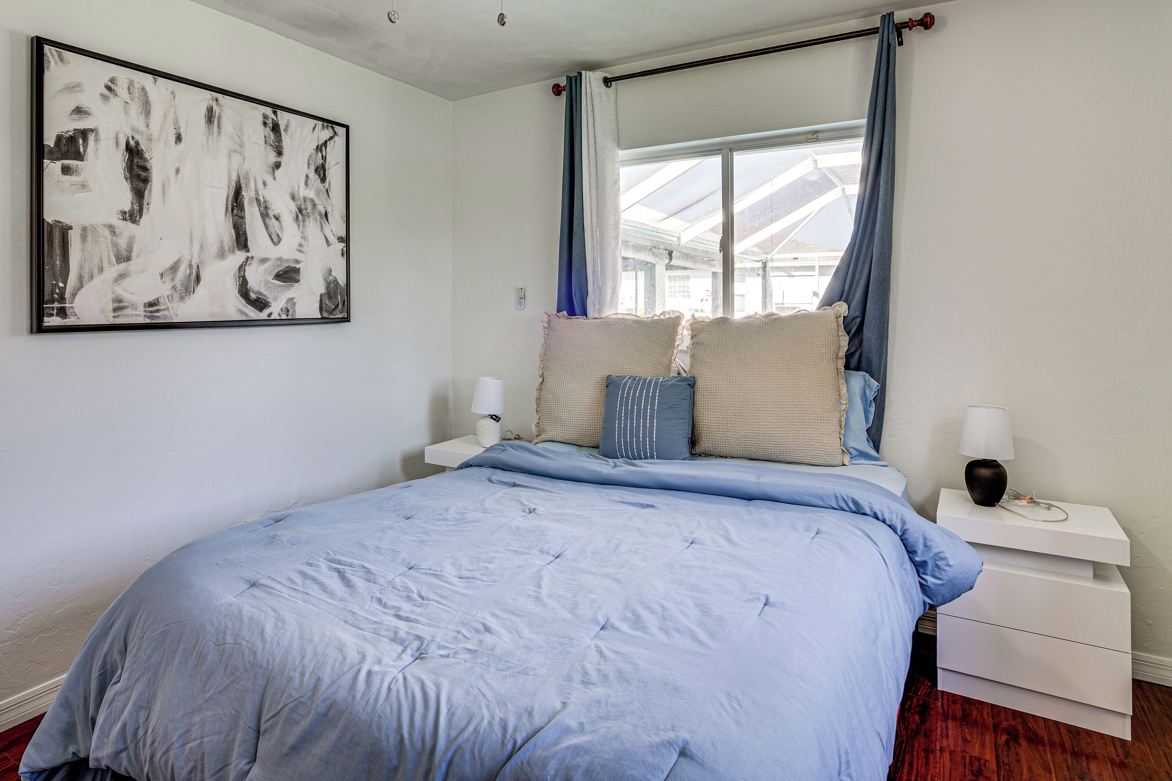 A bedroom with a large bed, a bedside table with a lamp, a wall-mounted artwork, and a window with curtains.