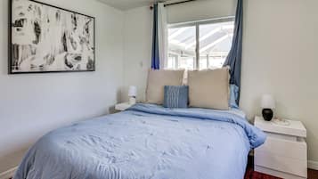 A bedroom with a large bed, a bedside table with a lamp, a wall-mounted artwork, and a window with curtains.