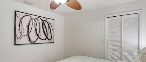 A bedroom with a bed, a ceiling fan, a wall-mounted artwork, and a closet with louvered doors.