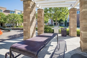 A covered outdoor seating area with a bench and a grill, surrounded by stone pillars and a view of residential buildings. - Walk to Train Station & More! Modern Compton Unit (Compton)