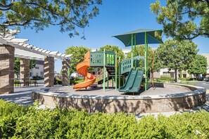 A playground with a slide and climbing structures, surrounded by a stone wall and greenery. - Walk to Train Station & More! Modern Compton Unit (Compton)