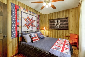 Ein Schlafzimmer mit Bett, Kissen, einer Steppdecke, einer Flagge, einem Deckenventilator und einem Stuhl.