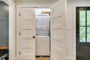 A laundry room with a washing machine and dryer, a door with a handle, and a window with a view of greenery. - Modern Farmhouse Between Mount Snow & Stratton! (Wardsboro)