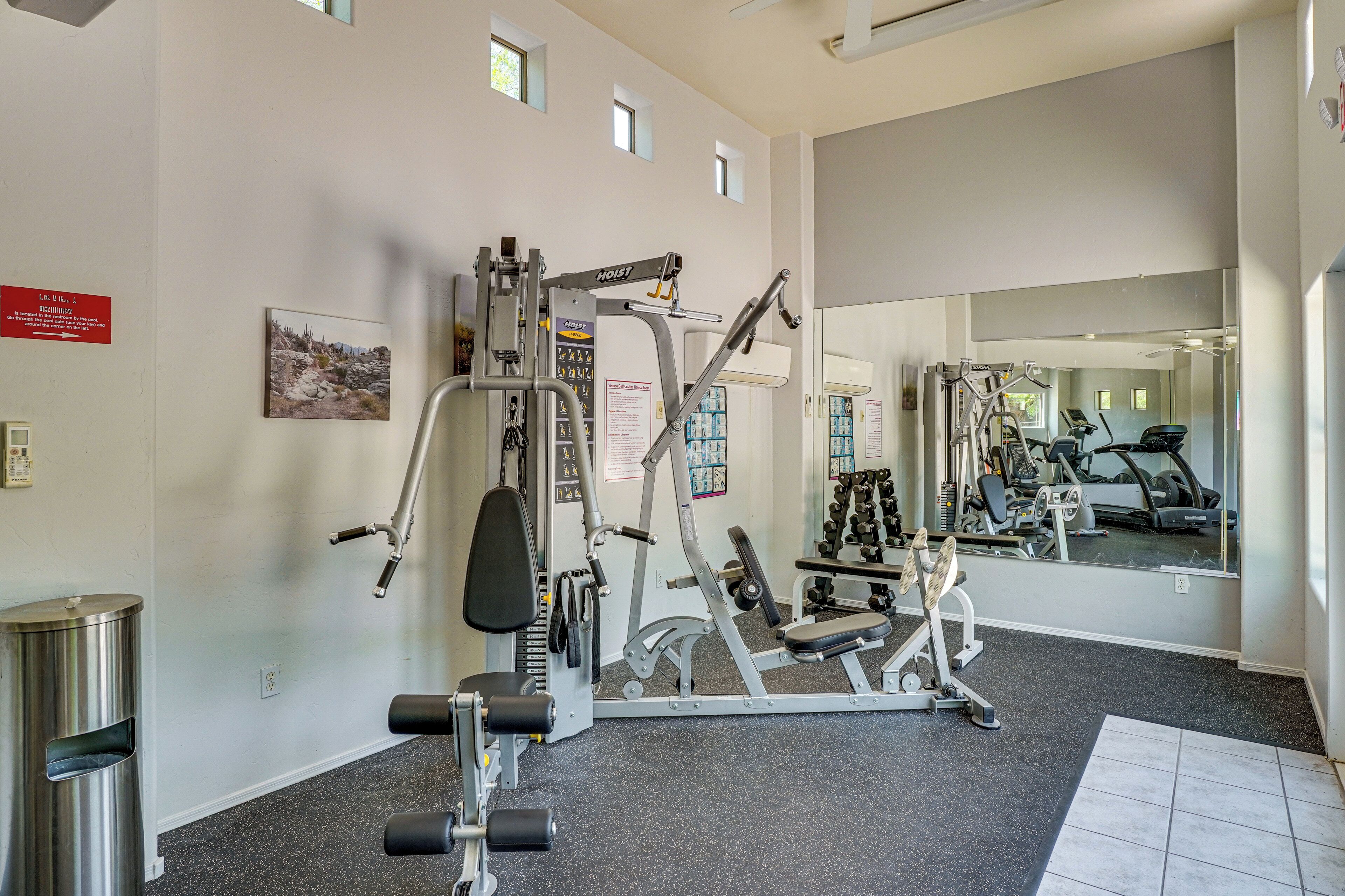 A gym with various exercise equipment, a mirror reflecting the equipment, and a sign indicating the location of the restrooms.