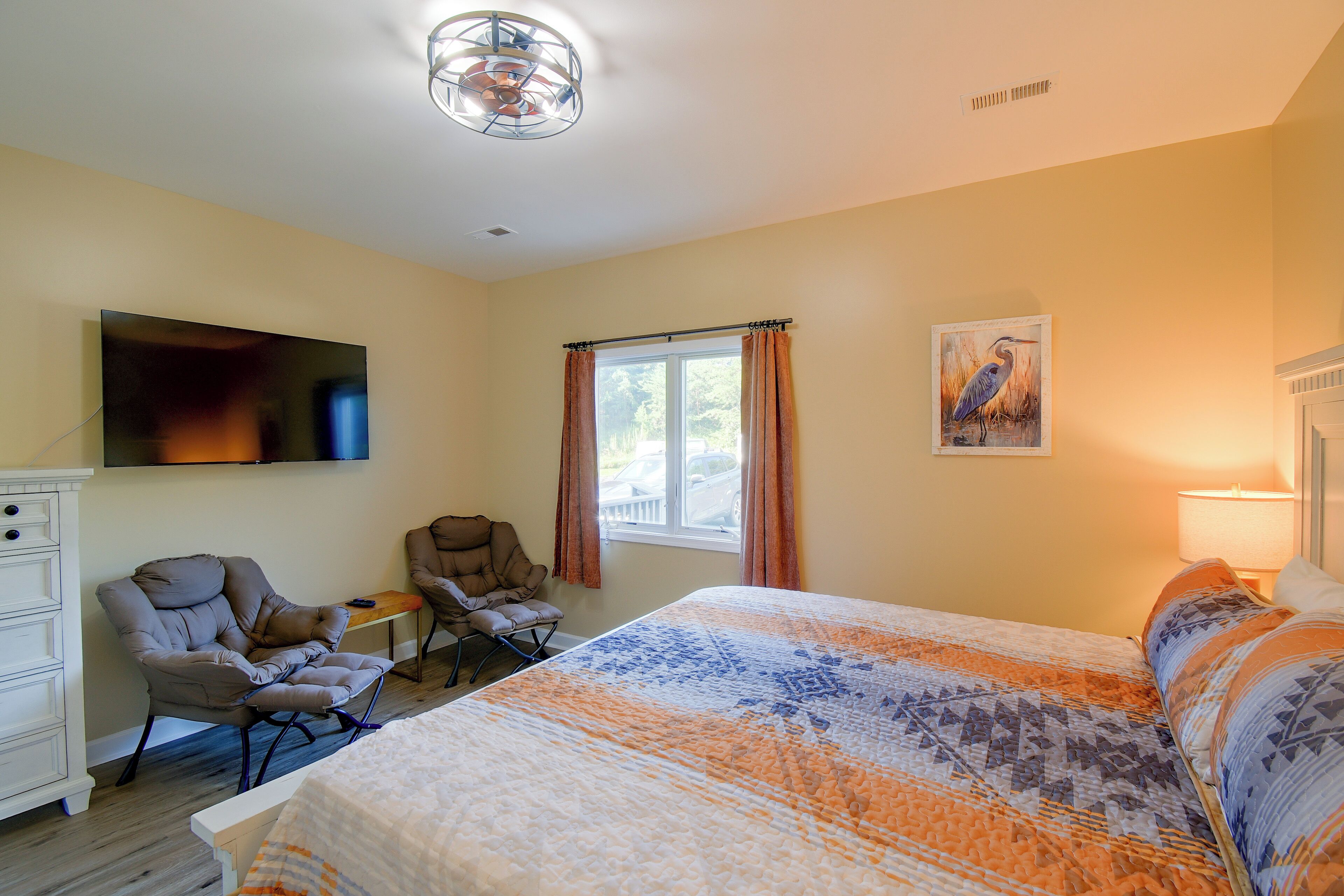 A bedroom with a bed, two chairs, a TV, a window with curtains, and a painting on the wall.