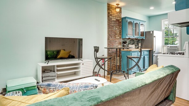 A compact living space with a sofa, a television, a kitchen area with blue cabinets, and a brick accent wall. - Walk to Hblr & Liberty State Park: Jersey City Apt (Jersey City)