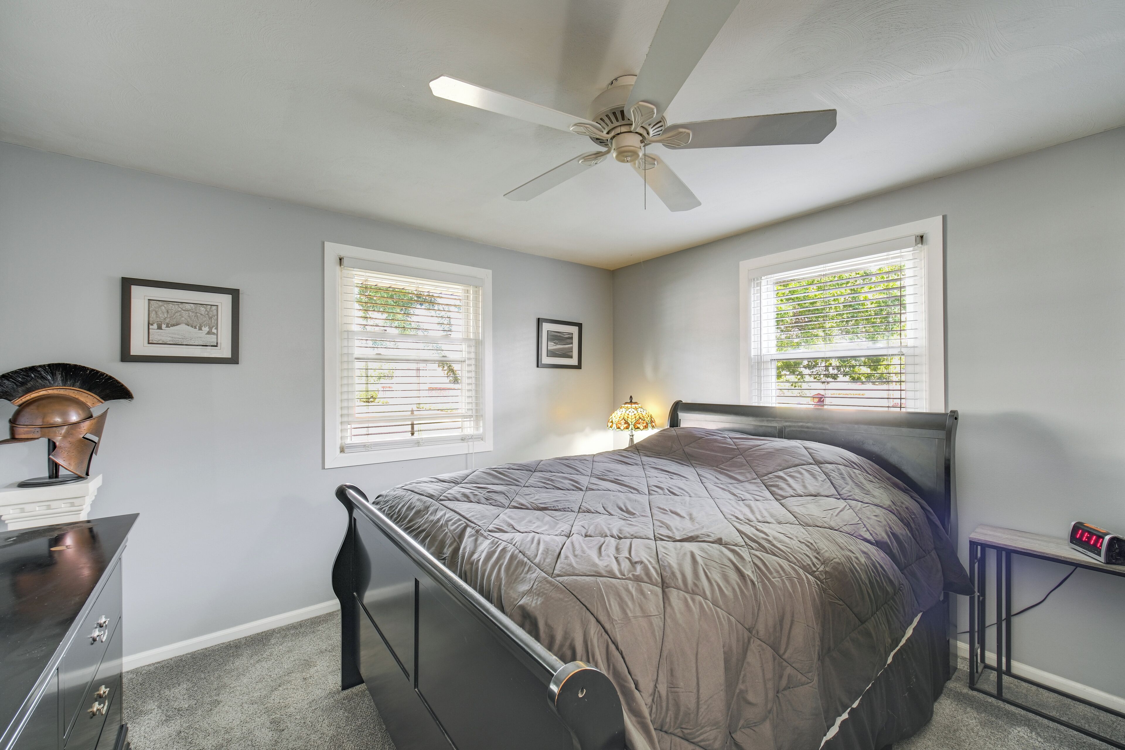 Une chambre à coucher comprenant un lit, un ventilateur de plafond, deux fenêtres avec stores, une table de chevet avec une lampe et une commode.