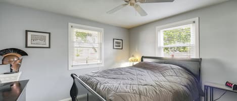 A bedroom with a bed, a ceiling fan, two windows with blinds, a nightstand with a lamp, and a dresser.