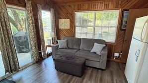 A wooden interior with a grey sectional sofa, a small ottoman, a refrigerator, and a window with blinds.