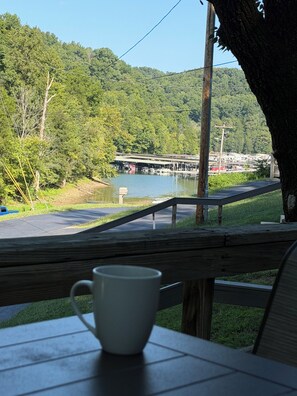 A wooden deck with a white mug, overlooking a lake with boats, a dock, and a forested hill.