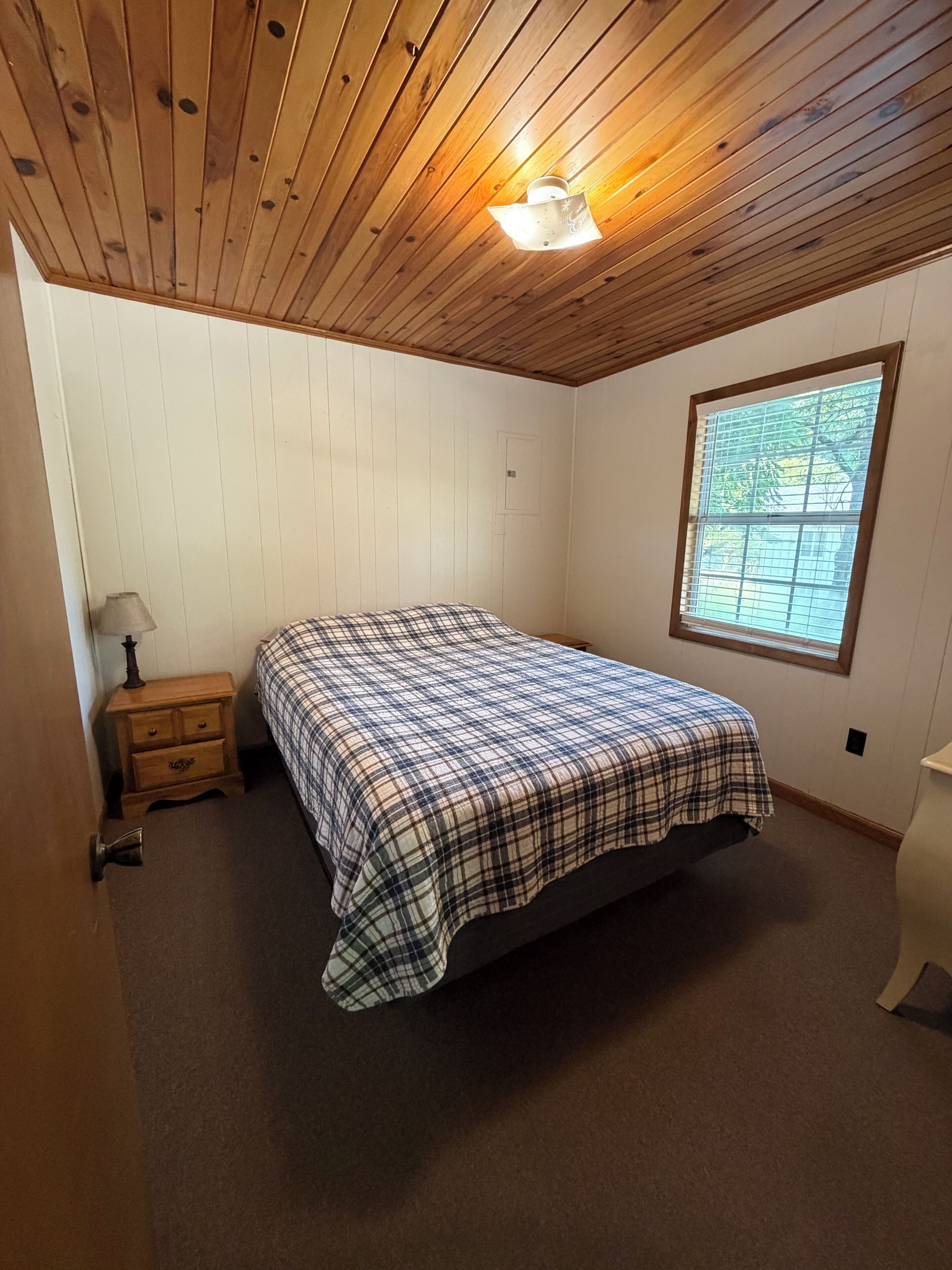 A bedroom with a wooden ceiling, a bed with a plaid bedspread, a nightstand, a window with blinds, and a door.
