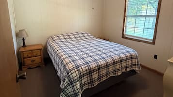 A bedroom with a wooden ceiling, a bed with a plaid bedspread, a nightstand, a window with blinds, and a door.