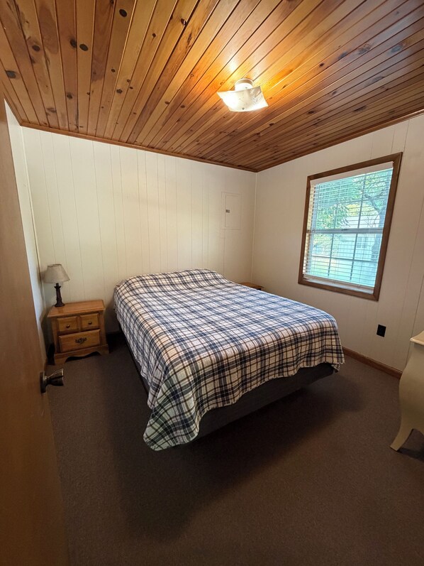 A bedroom with a wooden ceiling, a bed with a plaid bedspread, a nightstand, a window with blinds, and a door.