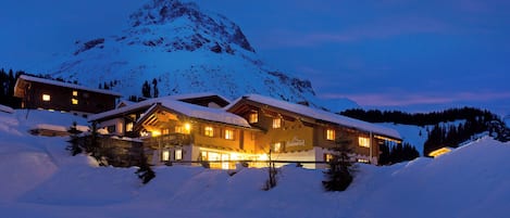 A snow-covered mountain lodge with multiple windows and a lit exterior at dusk.