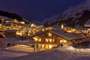 Vila montanhosa coberta de neve à noite, com edifícios iluminados e uma pousada destacada com varanda.