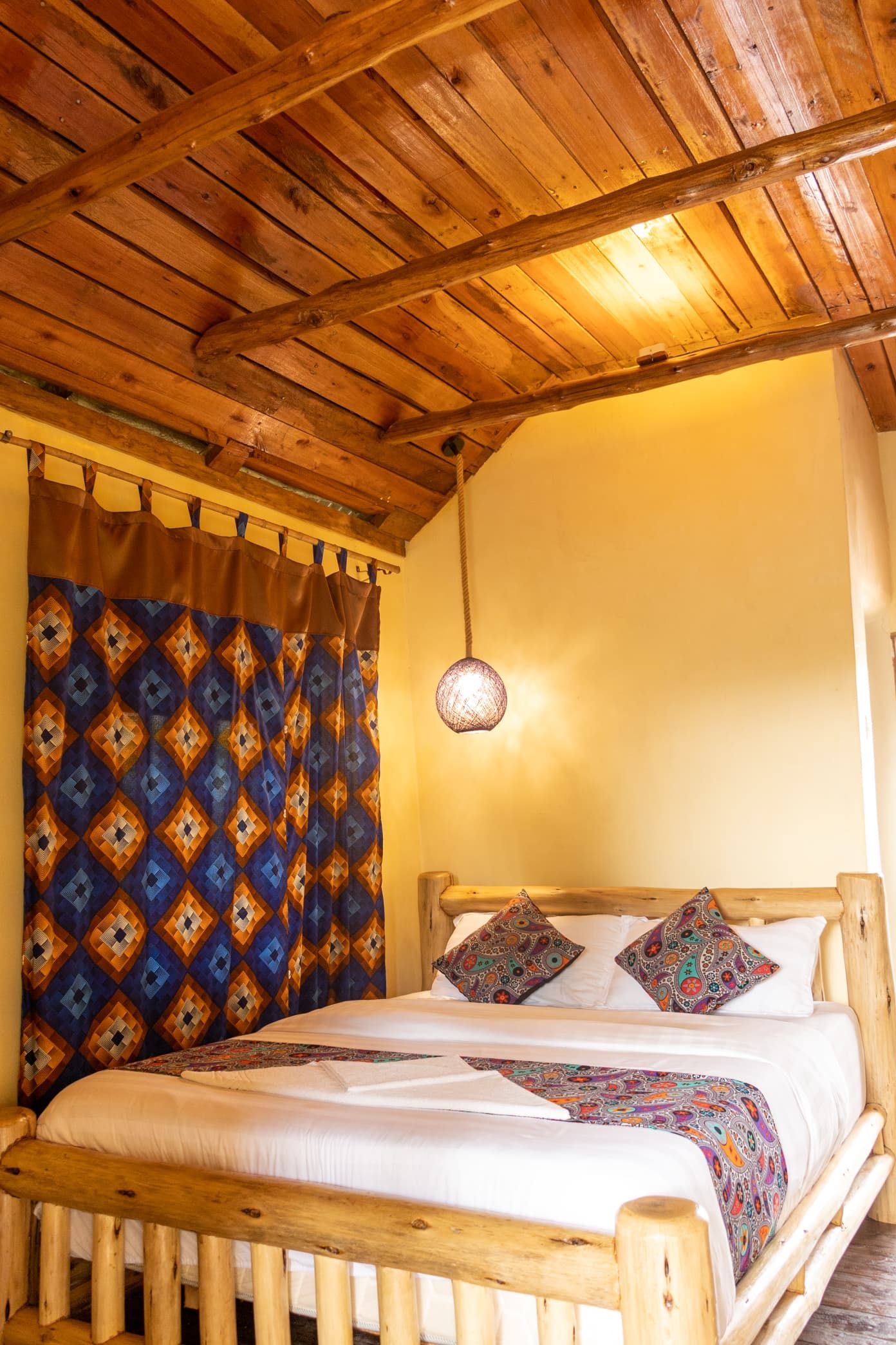 Ein hölzernes Bett mit einem gemusterten Bettbezug und Kissen, eine Pendelleuchte und eine Holzzieldecke.