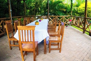 An outdoor dining area with a long table and multiple wooden chairs, surrounded by a wooden fence and lush greenery.