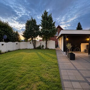 A residential backyard with a paved patio, a covered outdoor area, and a white fence.