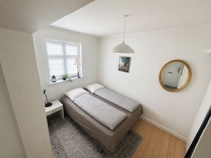 A bedroom with a single bed, a nightstand, a lamp, a window with blinds, a poster, and a mirror on the wall.