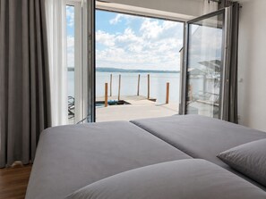 A bedroom with a large bed, a sliding glass door leading to a balcony with a view of the water and a dock.