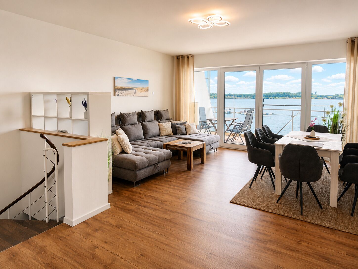 Ein modernes Wohnzimmer mit einer Couch, Esstisch und Stühlen, einer Treppe und einem großen Fenster mit Blick auf das Wasser.