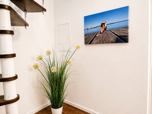 A white wall with a wooden floor, a white column, a potted plant with yellow flowers, and a framed photograph of a pier.