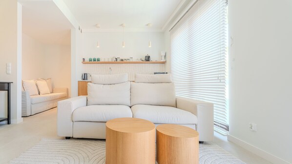 Un salon moderne avec un canapé, deux petites tables d’appoint en bois et, au fond, un coin cuisine.