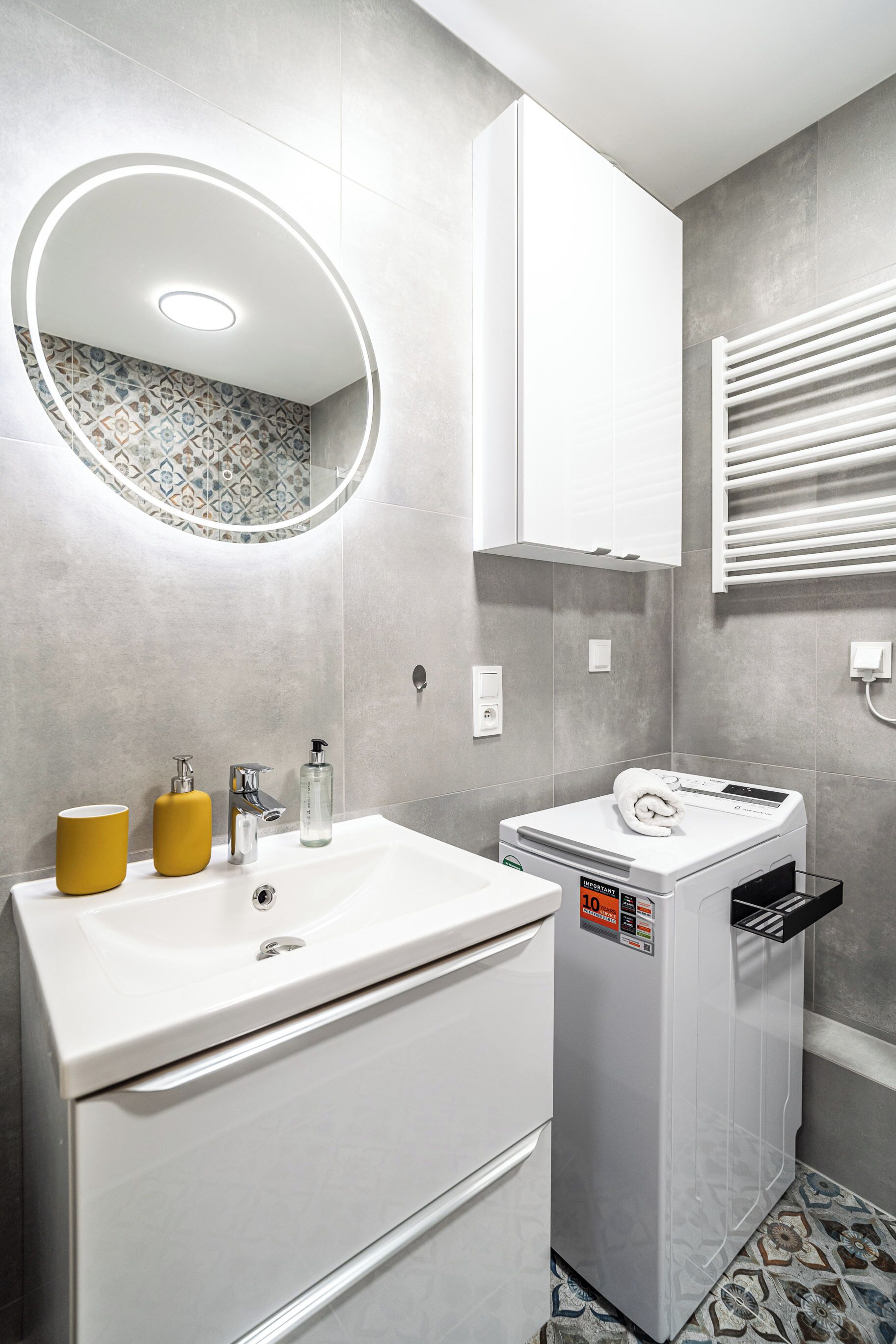 Une salle de bain moderne avec un lavabo blanc, des distributeurs de savon jaunes, un miroir avec un éclairage circulaire, un meuble blanc et un porte-serviettes.